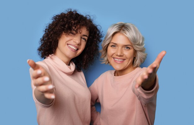 Cerrar foto de dos mujeres con cabello rizado sonriendo a la cámara e invitando a gesticular con las palmas en una pared azul del estudio