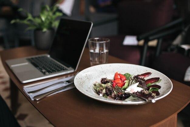 Cerrar foto de ensalada, portátil y vaso de agua y cuchillo con tenedor en la mesa en el restaurante