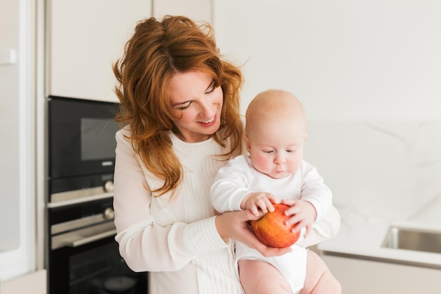 Cerrar foto de hermosa madre sonriente de pie en la cocina y sosteniendo a su lindo bebé y gran manzana roja en las manos