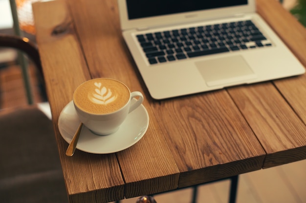 Cerrar foto de una mesa de madera con una taza de café en un plato. Portátil moderno en el fondo