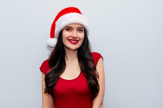 Cerrar foto de Navidad de mujer alegre con sonrisa radiante en vestido rojo y gorro de Papá Noel en el espacio gris.