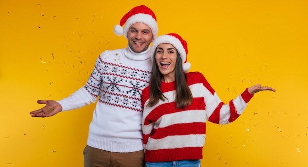 Cerrar foto de pareja joven emocionada en el amor en suéteres de invierno y gorros de Navidad Santa mientras se divierten y posando aislado sobre fondo amarillo