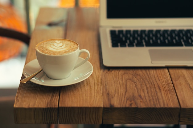 Cerrar foto de la superficie de una mesa de madera con un moderno portátil de pie en el fondo y una taza de café con leche en primer plano