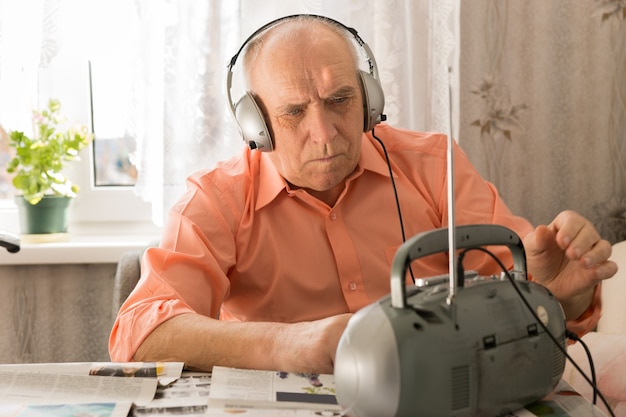 Cerrar hombre mayor serio escuchando noticias en el reproductor de casete seriamente blanco sentado en la sala de estar con periódicos.