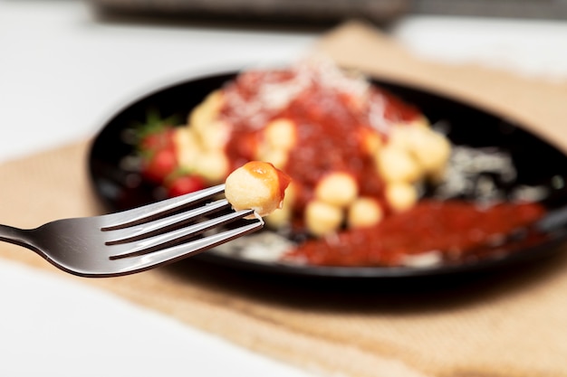 Foto cerrar en la horquilla con ñoquis y plato de ñoquis con salsa roja borrosa en el fondo