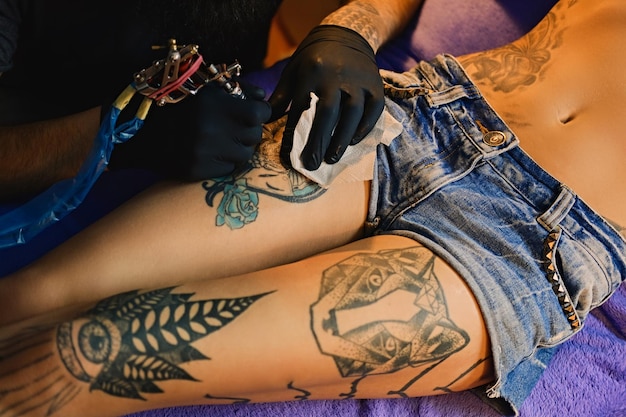 Cerrar la imagen del artista masculino del tatuaje barbudo hace un tatuaje en una pierna femenina.