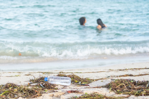 Cerrar imagen de botella de agua de plástico vacía en la playa sucia llena de algas, basura y desechos en la playa de arena sucia con gente en el mar