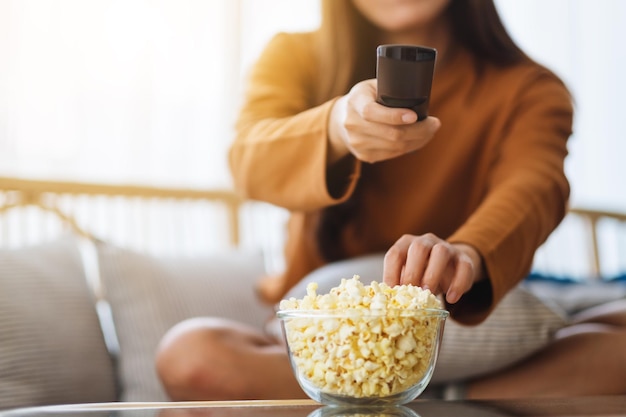 Cerrar imagen de una hermosa joven comiendo palomitas de maíz y buscando canal con control remoto para ver televisión mientras está sentado en el sofá en casa