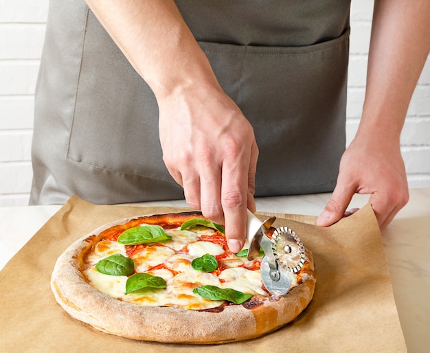 Cerrar la mano del chef panadero en uniforme gris cortando pizza en la cocina