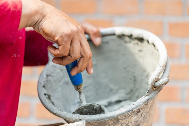 Foto cerrar la mano del trabajador sosteniendo la paleta y el balde con cemento fresco, albañil de construcción construyendo paredes con ladrillos, mortero y espátula