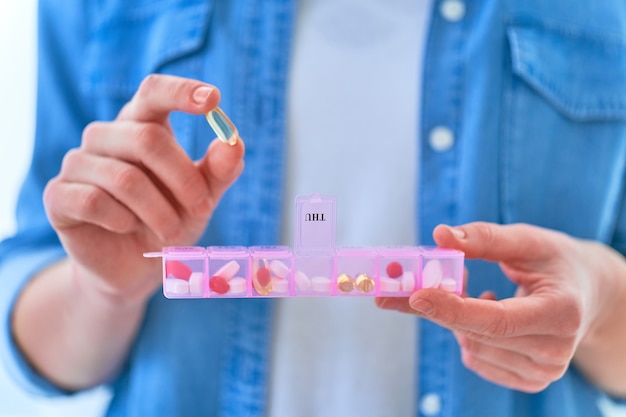 Foto cerrar las manos femeninas sosteniendo la caja de pastillas. mujer tomando medicamentos y suplementos dietéticos de omega 3 para la salud. cápsula blanda de aceite de pescado, vitamina d y c. dosis semanal de medicamento en un dispensador de píldoras