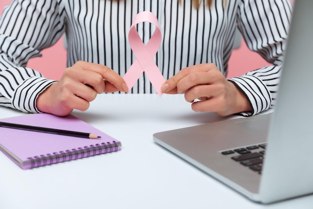 Cerrar las manos femeninas sosteniendo el símbolo de la cinta rosa del trabajador de la compañía de seguros de concientización sobre el cáncer de mama que trabaja en una computadora portátil haciendo notas Estudio interior aislado en fondo rosa