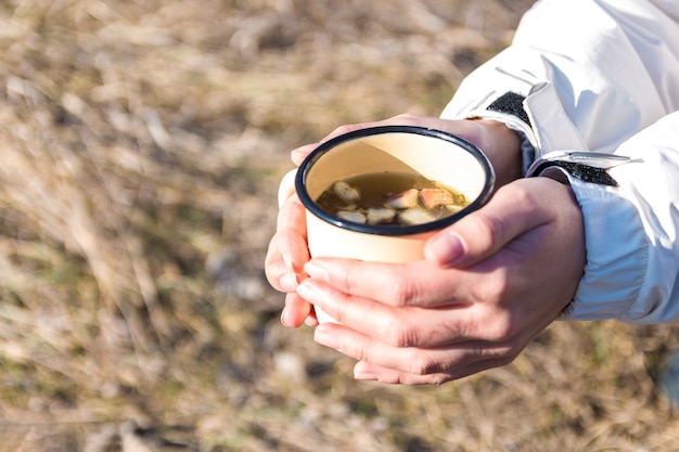 Cerrar las manos de una mujer excursionista en la celebración de la taza de metal con té de flores de campo