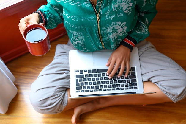 Cerrar las manos de la mujer sosteniendo una taza de té y escribiendo en la computadora portátil