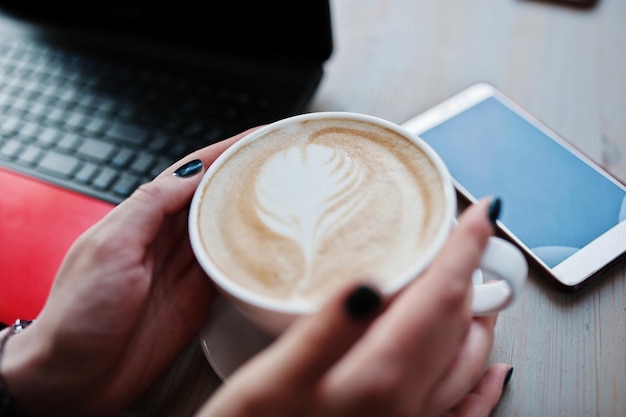 Cerrar manos de niña con taza de capuchino fondo rojo portátil y teléfono móvil