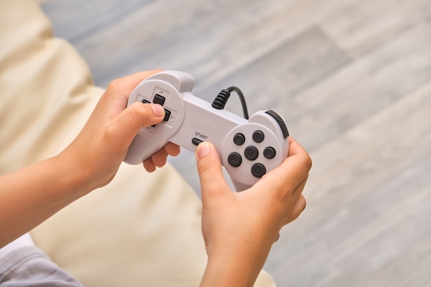 Cerrar las manos del niño sosteniendo un controlador de joystick mientras juega videojuegos en casa, sentado en el sofá en casa