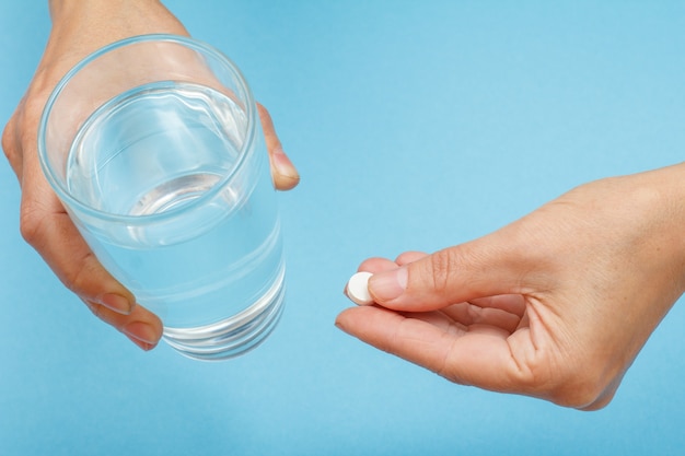 Cerrar las manos de la paciente con un vaso de agua en una mano y una pastilla blanca en la otra. Vista superior con fondo azul y espacio de copia