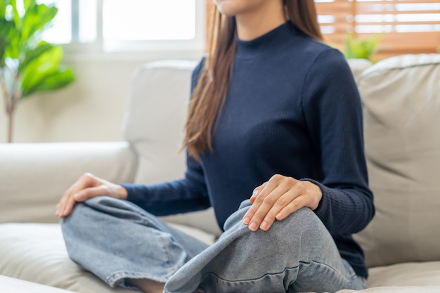 Cerrar las manos de la postura tranquila mujer joven asiática práctica sentada meditando en posición de loto en el sofá en casa ejercicio de meditación para el bienestar cuidado saludable Relajación ocio personas