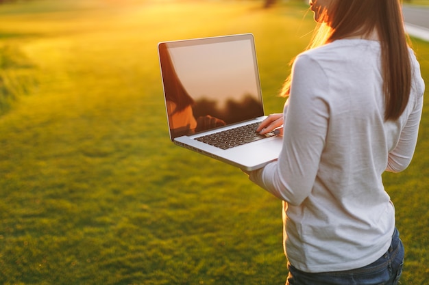 Cerrar las manos en el teclado. Mujer que trabaja en la computadora portátil con pantalla vacía en blanco negro para copiar el espacio en el parque en el césped de hierba verde sol al aire libre. Oficina móvil. Concepto de negocio autónomo.