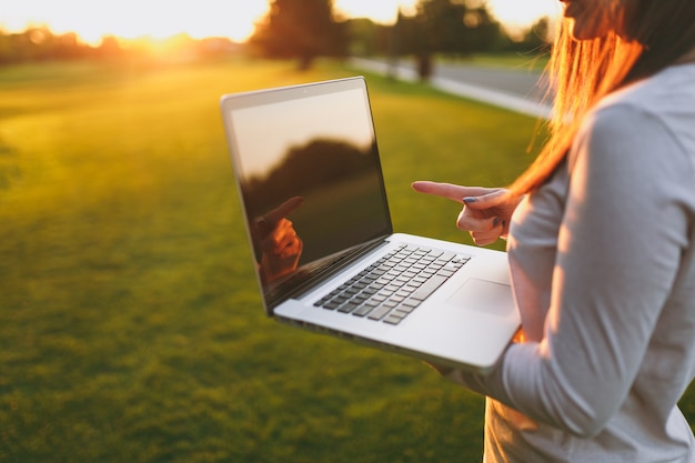 Cerrar las manos en el teclado. Mujer que trabaja en la computadora portátil con pantalla vacía en blanco negro para copiar el espacio en el parque en el césped de hierba verde sol al aire libre. Oficina móvil. Concepto de negocio autónomo.