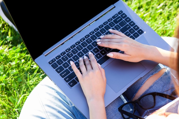 Cerrar las manos en el teclado. Mujer que trabaja en la computadora portátil con pantalla vacía en blanco negro para copiar el espacio en el parque en el césped de hierba verde sol al aire libre.