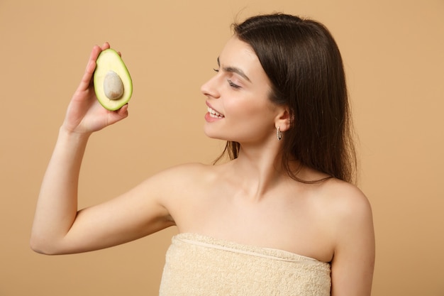 Cerrar morena mujer medio desnuda con piel perfecta maquillaje desnudo sostiene aguacate aislado en la pared de color beige pastel