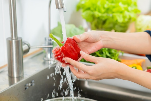 Cerrar mujer asiática joven ama de llaves lavando pimiento rojo pimentón verduras con agua salpicada en un recipiente de agua en el fregadero en la cocina preparando ensalada fresca cocinando comida Gente de alimentos saludables