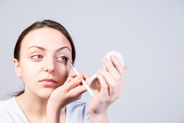 Cerrar mujer joven aplicar maquillaje delineador de ojos mientras mira el pequeño espejo de un kit de base sobre un fondo gris.