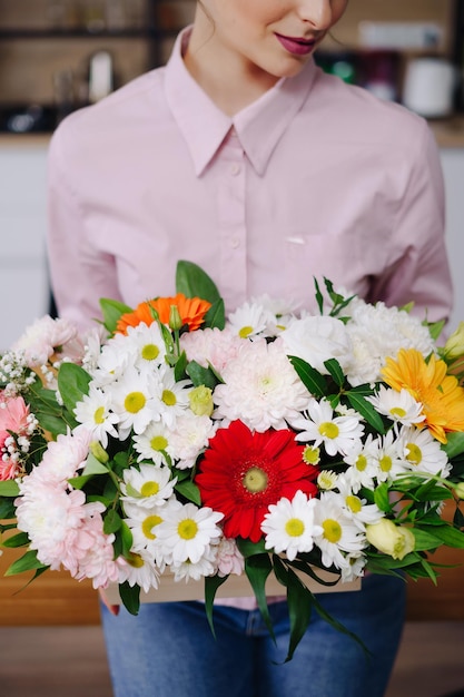 Cerrar mujer sosteniendo una composición de flores posando en la cocina blanca en casa con una computadora portátil Floreria presentando una canasta con flores de colores