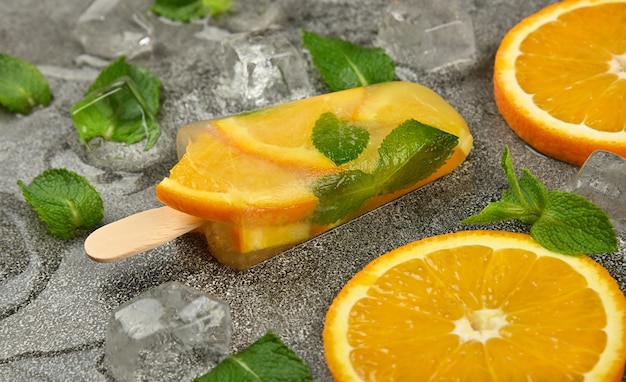 Cerrar una paleta de helado de fruta con rodajas de naranja fresca, hojas de menta verde y cubitos de hielo en la superficie de la mesa gris, vista de ángulo alto