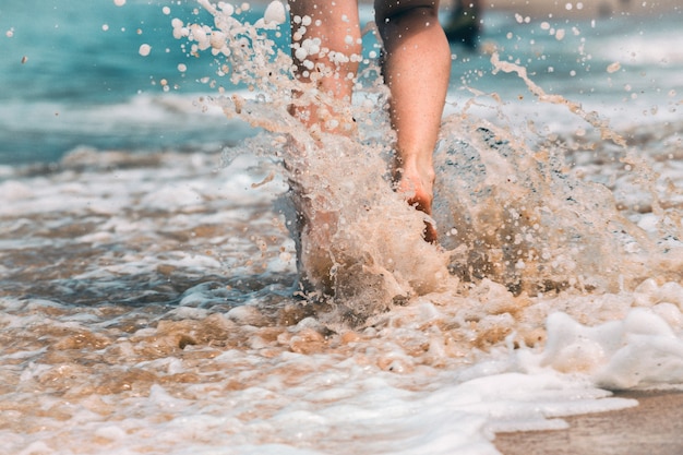 Cerrar la pierna de una mujer joven caminando por la ola de agua de mar y arena en la playa de verano. Concepto de viaje.