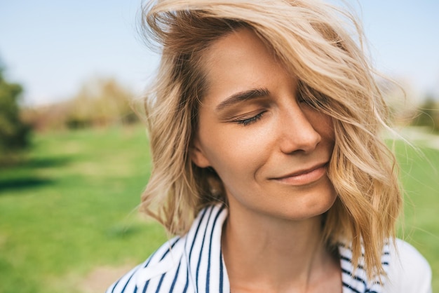 Cerrar el retrato al aire libre de una mujer bonita con el pelo rubio que sopla y los ojos cerrados disfrutar del clima Cerrar tiro de una hermosa joven caucásica sonriendo con el pelo ventoso en el parque Concepto de personas