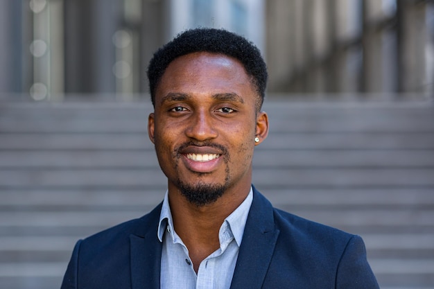 Cerrar retrato feliz hombre de negocios afroamericano mirando a cámara exterior. Cara de sonrisa apuesto hombre de negocios negro en traje formal en la calle de la ciudad urbana moderna fondo borroso al aire libre closeup