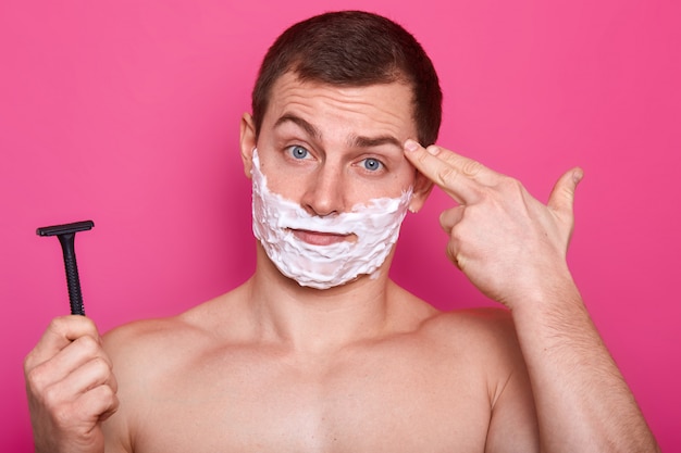 Cerrar retrato de hombre guapo con espuma de afeitar en la cara, mantiene la maquinilla de afeitar en la mano lista para afeitarse
