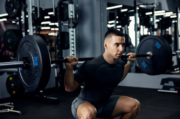 Cerrar el retrato lateral de un entrenador de fitness masculino en ropa deportiva gris haciendo sentadillas con una barra en un gimnasio