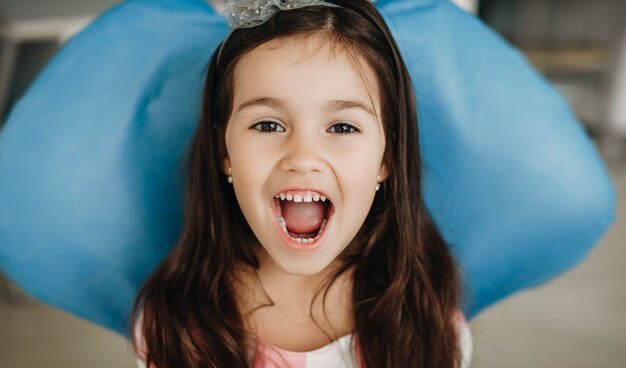 Cerrar el retrato de una linda niña sentada en un asiento de estomatología mirando a la cámara mostrar los dientes después de la cirugía de dientes en una estomatología pediátrica.