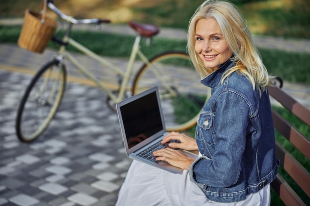 Cerrar retrato de mujer alegre feliz en chaqueta de mezclilla mirando a la cámara de fotos mientras está sentado en el banco de madera frente a la bicicleta