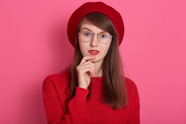 Cerrar retrato de mujer joven mantiene los dedos delanteros cerca del labio inferior, estar en alto espíritu, vistiendo boina roja, suéter y gafas