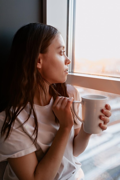 Cerrar el retrato de perfil de una adorable y encantadora dama con café matutino mirando por la ventana a la luz del sol Retrato interior de una joven mujer elegante en casa