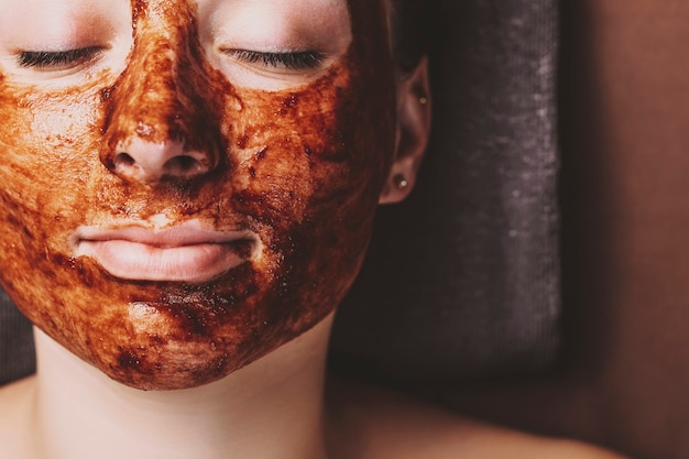 Foto cerrar el rostro femenino apoyado en una cama de spa con los ojos cerrados con máscara de chocolate contra el acné en el centro de spa.