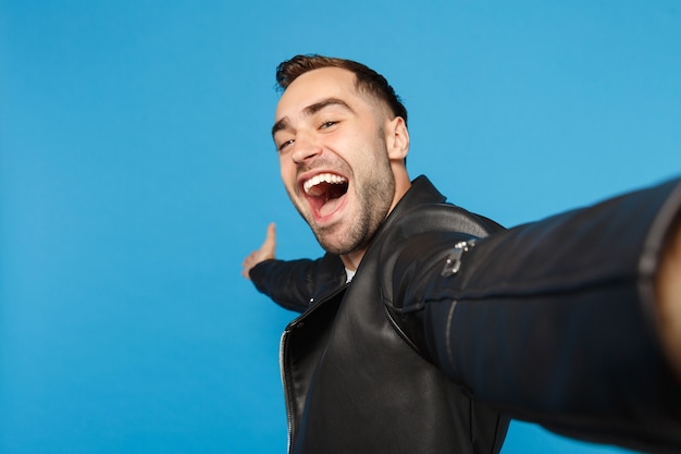 Cerrar selfie de elegante joven sin afeitar en chaqueta de cuero negro camiseta blanca mirando cámara aislada en retrato de estudio de fondo de pared azul. Concepto de emociones sinceras de personas. Simulacros de espacio de copia