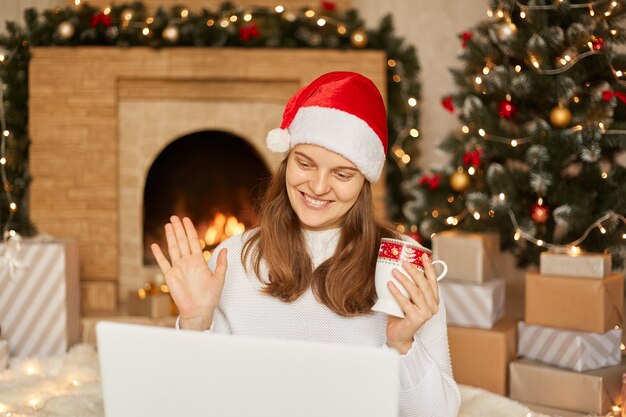 Cerrar sonriente mujer europea usando una computadora portátil, mirando la pantalla, saludando con la mano, saludando, charlando en línea con alguien, haciendo videollamadas en el árbol de Navidad y la chimenea