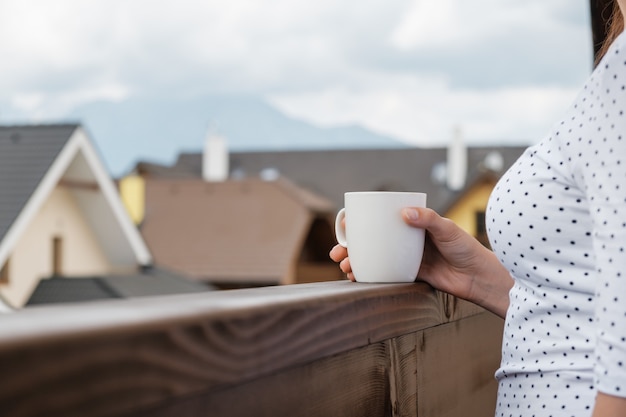 Cerrar una taza blanca en una mano de mujer en el balcón de madera con montañas y techo de la casa. mañana de vacaciones