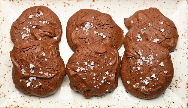 Cerrar varias galletas de chocolate brownie redondas recién horneadas con sal en escamas en un plato de porcelana blanca, vista superior elevada, directamente encima