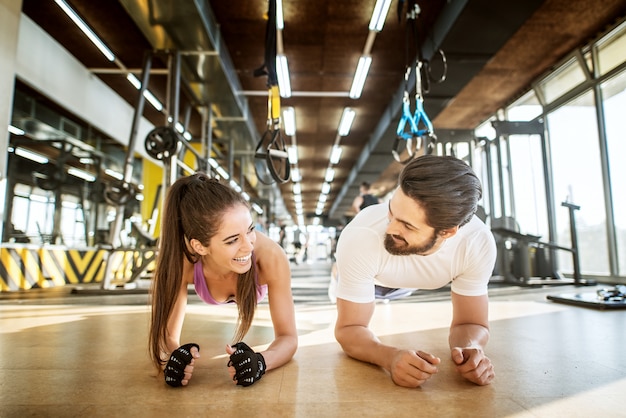 Cerrar la vista frontal de la adorable chica atractiva sonriente haciendo plancha mientras está de pie con su entrenador personal en el soleado gimnasio mientras se miran.