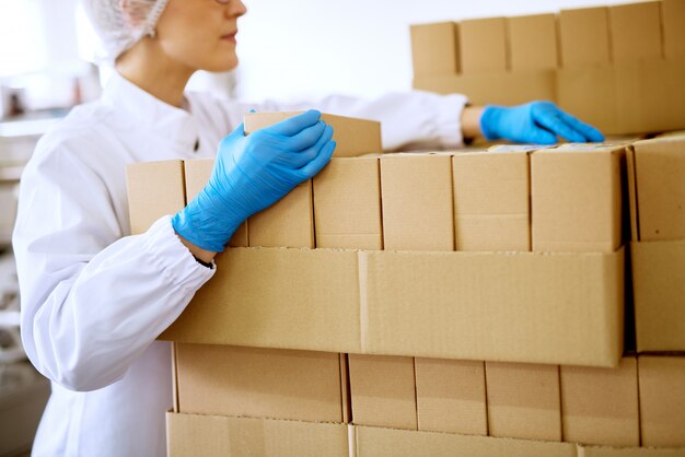 Cerrar vista de una joven trabajadora enfocada en paños estériles y guantes de goma azul contando cajas apiladas en una sala de almacenamiento de la fábrica.