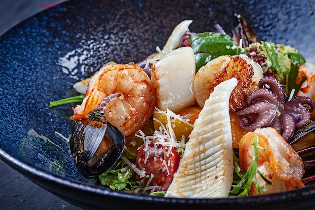 Cerrar vista sobre ensalada con mariscos servidos en un recipiente oscuro. Fotografía de alimentos para anuncios o recetas. Copia espacio Ensalada tibia con camarones, calamares, vieiras, pulpo en un tazón. Almuerzo merienda.