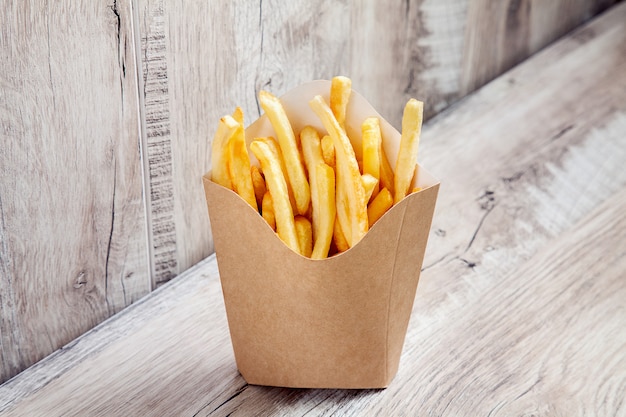 Cerrar vista sobre patatas patatas fritas en caja de paquete de cartón aislado sobre fondo de madera. Concepto de comida rápida simulacro. Cartón de papel kraft o artesanal en blanco con papas fritas