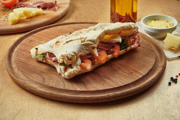 Cerrar vista sobre sabroso panuozzo italiano tradicional sandwich - tortilla al horno con tomates, mozzarella derretida, jamón y rúcula sobre una tabla de madera. comida de la calle