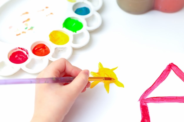 Cerrar vista superior de la mano del niño con pincel dibujando el sol sobre la casa en papel blanco.